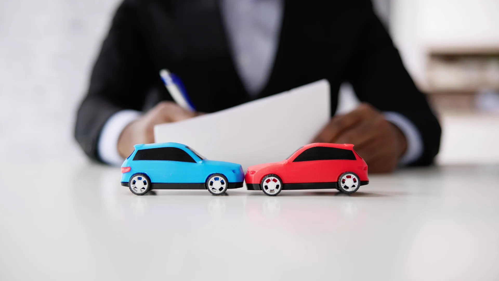 Close-up of two toy cars in a crash position with a person reviewing documents in the background, symbolizing an auto accident claim or insurance review.