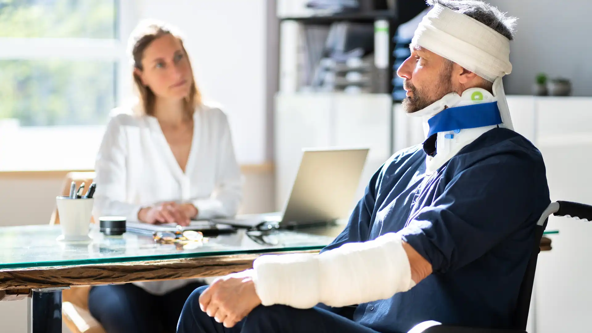 Injured man with arm cast and head bandage speaking with a professional woman at a desk.
