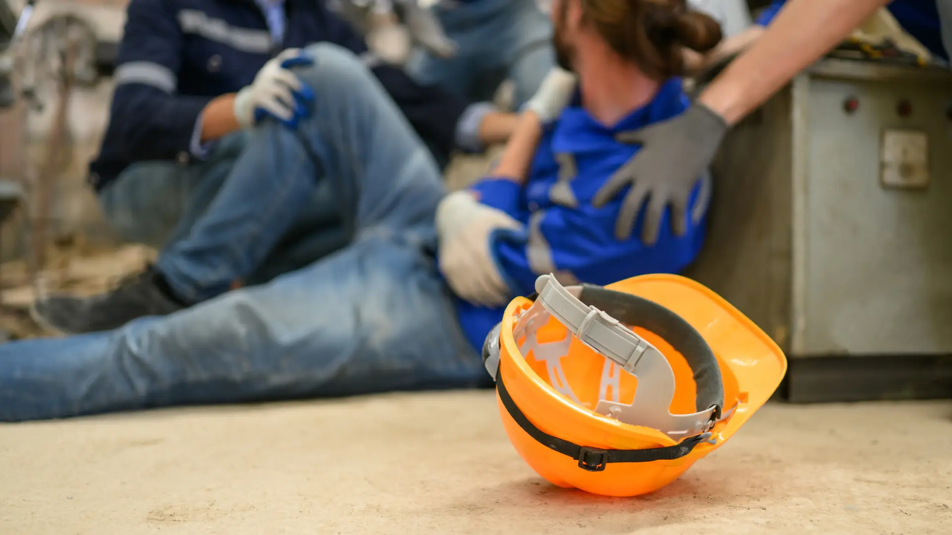 "Injured construction worker sitting on the ground with others assisting, safety helmet in foreground