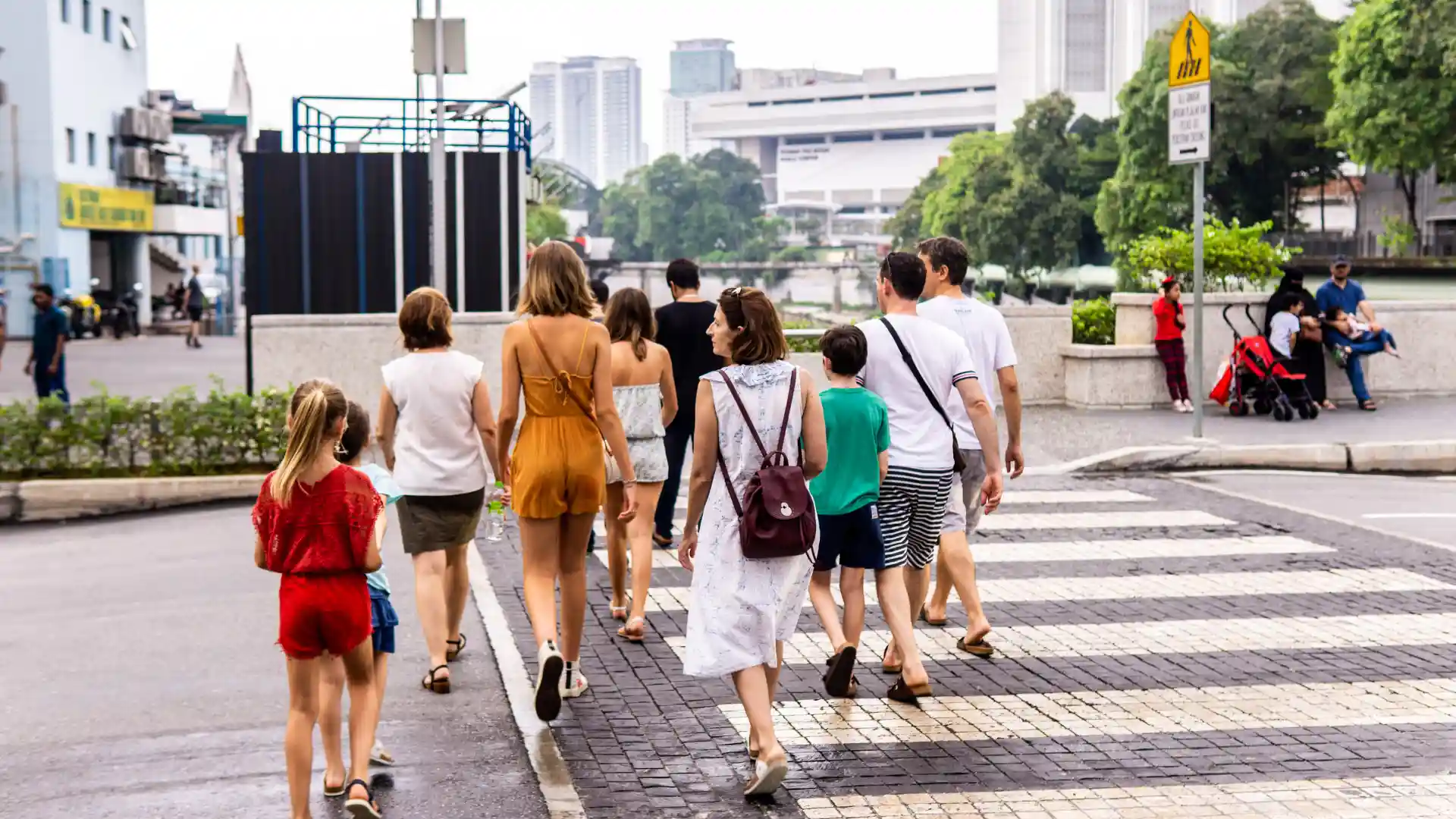 Group of people walking across a city crosswalk on a bright day, including adults and children.